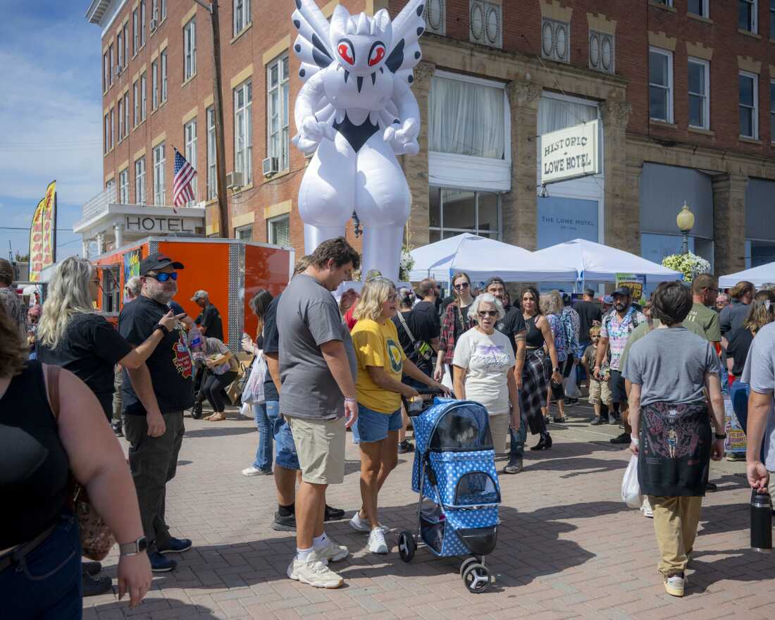 With the Lowe Hotel as a backdrop, attendees of the Mothman Festival 2025 congregate along the historic Main Street. There is a white inflatable Mothman-like figure towering over the crowd.