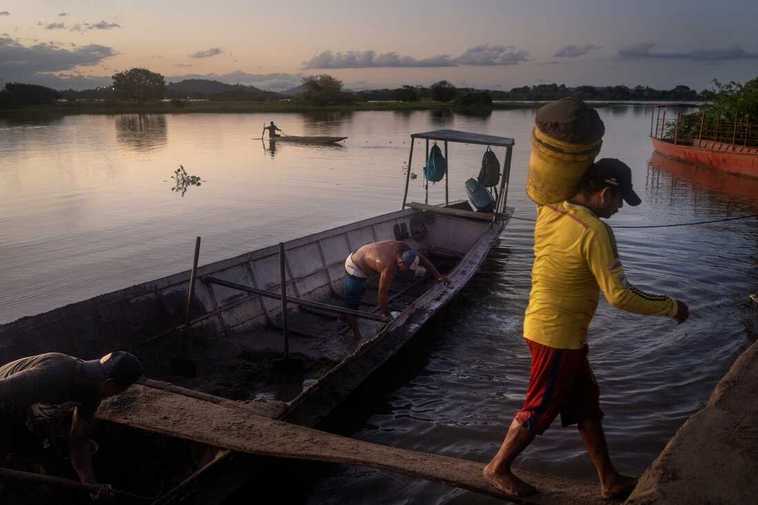 Laborers unload sand at the El Cesar dock in El Banco, Colombia, on August 8, 2022. A fishing boat is visible returning to the dock in the background. El Banco and its surroundings along the Magdalena River are widely regarded as the birthplace of cumbia. The National Cumbia Festival, held in El Banco Magdalena, was established in 1968 to honor the legendary composer José Benito Barros Palomino.