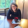 A smiling woman sits at a desk with arms crossed. There's a plant and a ceramic mug and papers on the desk.