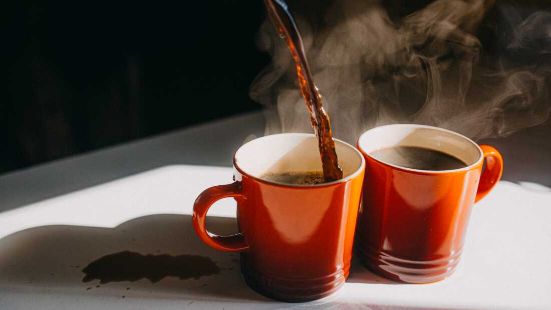 A photograph shows hot coffee being poured simultaneously into two burnt orange coffee mugs on a white countertop. Steam rises from the mugs.