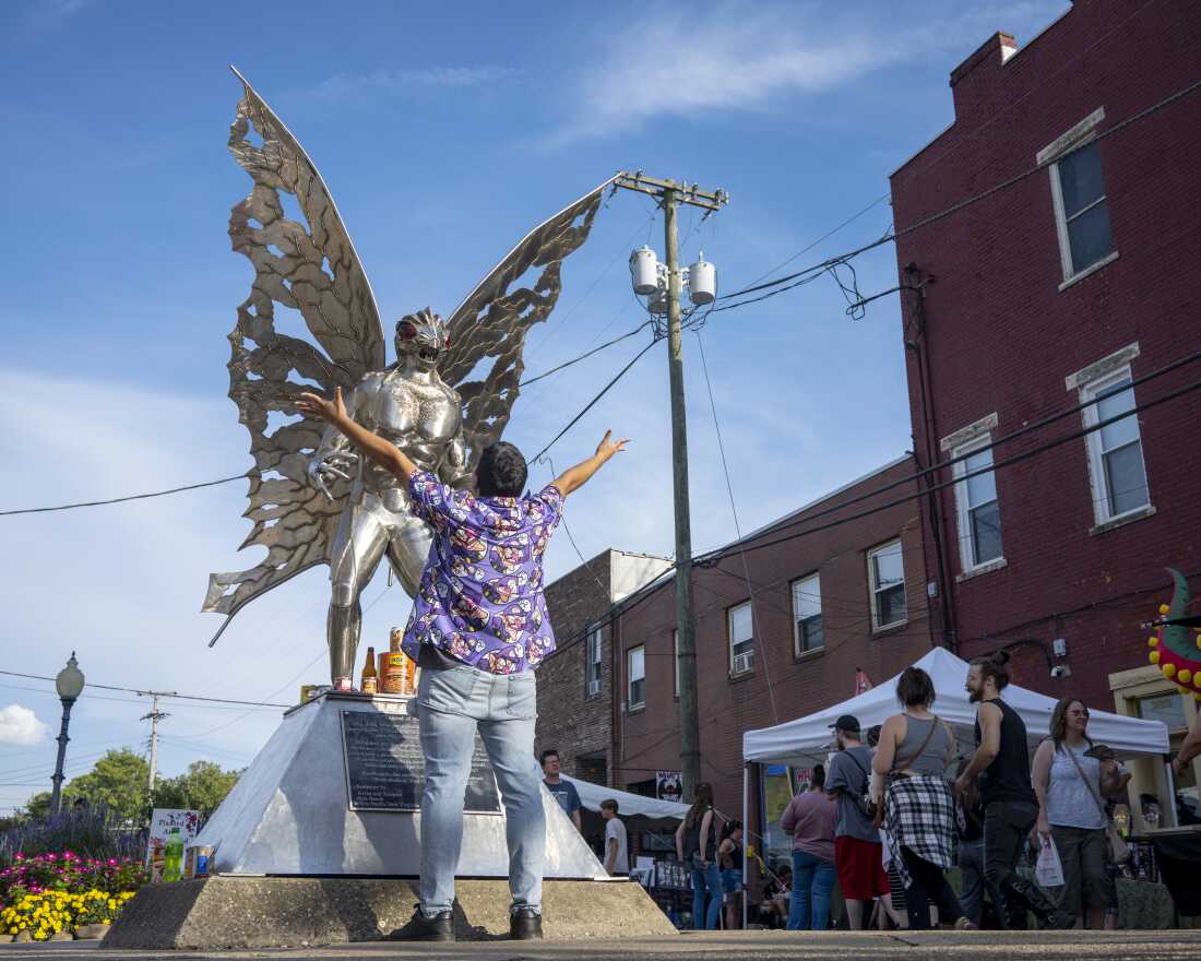 Michael Negron of Norwalk, Conn., wearing jeans and a flowered purple shirt, raises his arms as he faces the silver Mothman sculpture in Point Pleasant, W.Va.