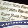 Close-up of a blue Social Security card with U.S. Treasury checks partially visible behind it, featuring the Statue of Liberty torch emblem.